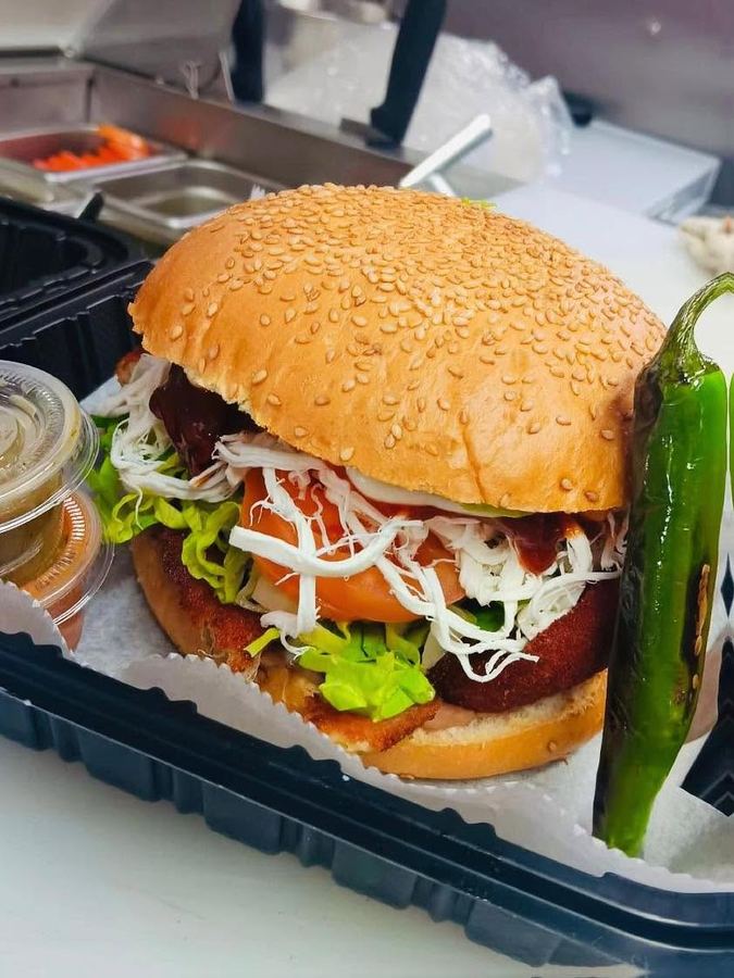 A cemita on a sesame bun stacked with breaded milanesa, stringy Oaxaca cheese, lettuce and tomato, with a roasted jalapeño alongside.