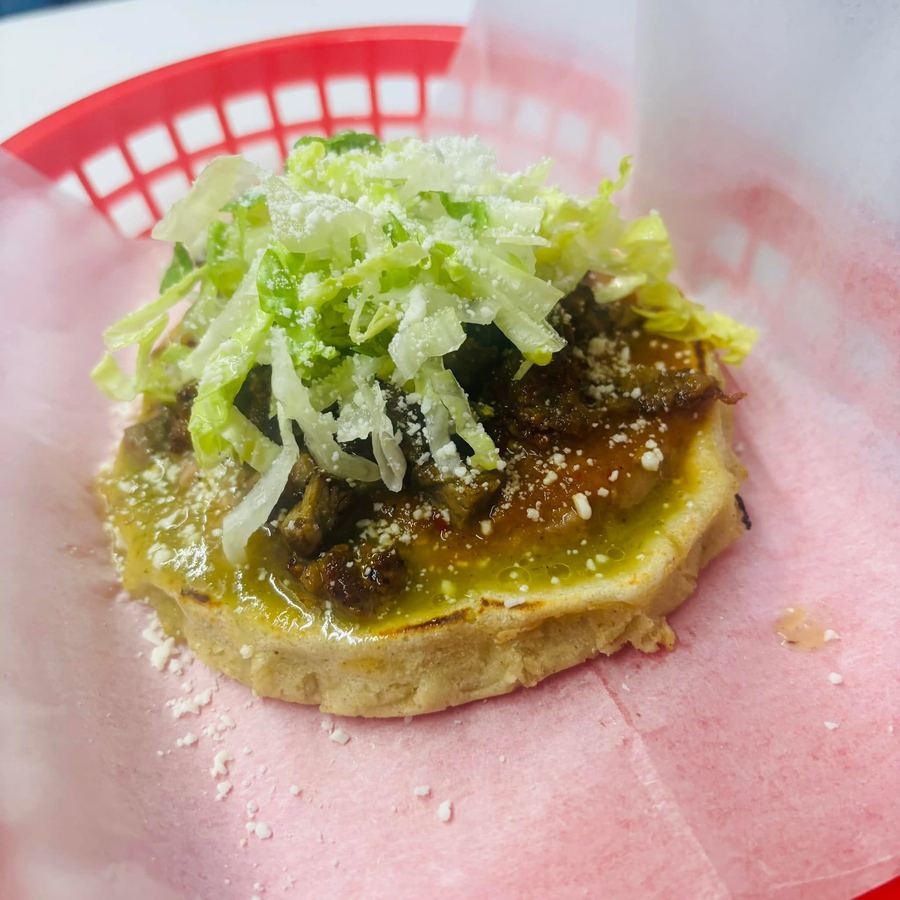 A hand-pressed masa base in a red basket, topped with salsa verde, meat, shredded lettuce and crumbled queso.