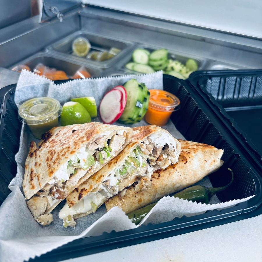 A chicken quesadilla cut into wedges in a takeout tray, with limes, radish, cucumber and salsas on the side.