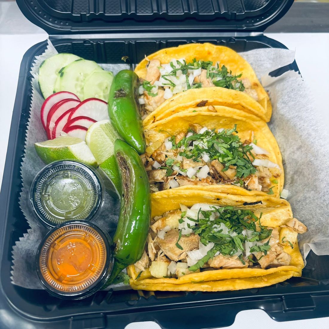 An order of tacos topped with onion and cilantro, served with limes, radish, cucumber, roasted jalapeños and two salsas.